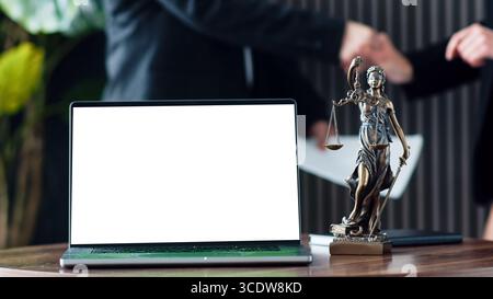 Statue de la justice se tient à côté de l'ordinateur portable avec écran blanc sur le bureau en bois, comme deux avocats se serrent la main dans le fond, représentant le partenariat juridique an Banque D'Images