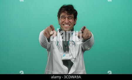 Médecin hispanique avec des lunettes et un stéthoscope pointant vers la caméra sur fond vert éclatant, souriant avec confiance. Banque D'Images