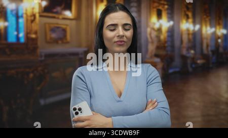 Femme se tient soigneusement à l'intérieur d'un musée orné portant une chemise bleue avec ses bras croisés et tenant un smartphone au milieu d'œuvres d'art faiblement éclairées. Banque D'Images