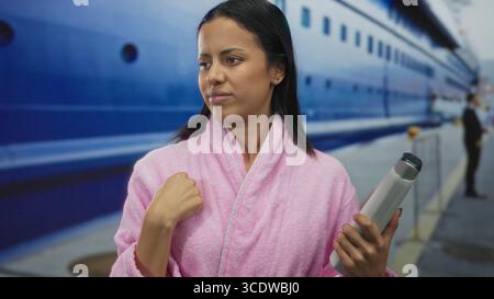 Femme en peignoir rose tenant une bouteille, se tient à l'extérieur près d'un navire amarré, incarnant une ambiance balnéaire sereine avec une toile de fond maritime. Banque D'Images