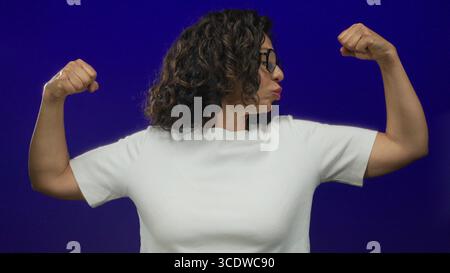 Femme hispanique d'âge moyen fléchissant en toute confiance ses muscles contre un mur de fond bleu isolé, mettant en valeur la force et l'autonomisation dans son décontracté Banque D'Images