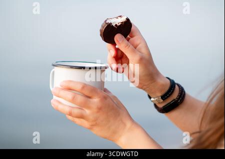 Gros plan détaillé des mains d'une femme tenant une tasse de boisson chaude et un bonbon au chocolat, capturant un moment confortable et indulgent Banque D'Images