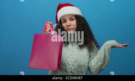 Femme portant le chapeau de père noël et le pull tient le sac à provisions rose avec l'expression confuse devant le fond bleu indiquant le dilemme du shopping de vacances. Banque D'Images