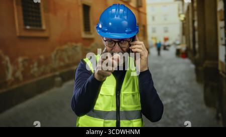Homme en casque bleu et gilet jaune, parlant au téléphone dans un environnement urbain de rue, souriant et pointant, travailleur de la construction d'âge moyen à l'extérieur. Banque D'Images