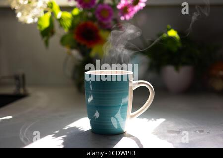 Une tasse de café fumant se trouve sur un comptoir de cuisine, éclairé par la lumière du soleil. Des fleurs colorées décorent le fond, créant une atmosphère chaleureuse et accueillante remplie d'énergie matinale. Banque D'Images