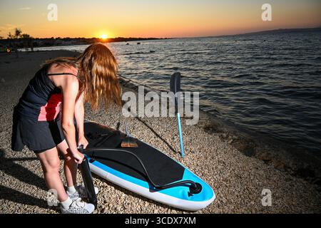 Sportive gonflant sup board sur la plage de galets au coucher du soleil, se préparant pour le paddleboard. Banque D'Images