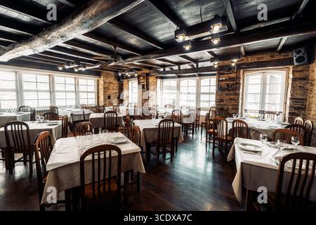 Plan grand angle discret d'un intérieur de restaurant rustique confortable avec poutres en bois, murs en pierre, tables soigneusement agencées recouvertes de linge blanc ; grand bri Banque D'Images