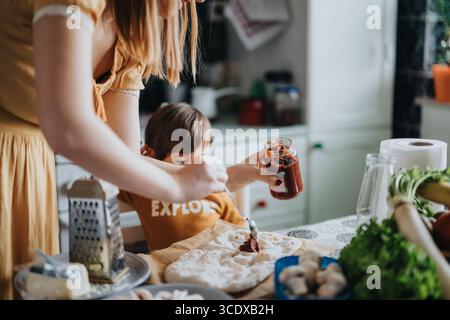 Mère et enfant préparant une pizza maison avec de la sauce tomate dans une atmosphère de cuisine confortable Banque D'Images