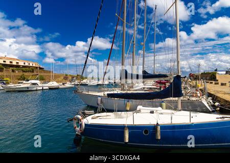 Voiliers amarrés et yachts dans le port paisible de Stintino, en Sardaigne, sous un ciel bleu vibrant avec des nuages blancs moelleux. Banque D'Images