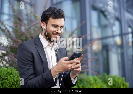 Un homme professionnel bien habillé en costume s'engage avec son smartphone et ses écouteurs à l'extérieur. Banque D'Images