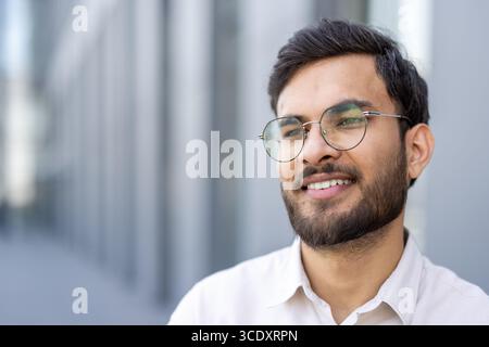Un jeune homme sourit en toute confiance en portant des lunettes, exsudant bonheur et détente dans un environnement urbain. Banque D'Images