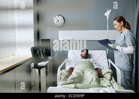 Infirmière caucasienne tenant une planche à pince avec dossier médical debout dans un service hospitalier posant des questions à un patient noir Banque D'Images