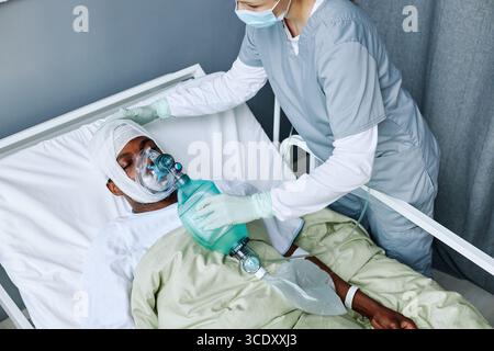 Vue en grand angle d'une infirmière caucasienne travaillant dans un hôpital moderne en ventilation artificielle pour un jeune homme noir traumatisé crânien Banque D'Images