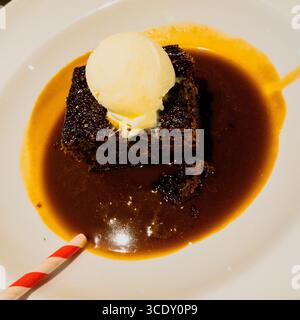 Pudding au caramel collant dans un bol blanc avec de la crème glacée à la vanille et une paille rayée Banque D'Images