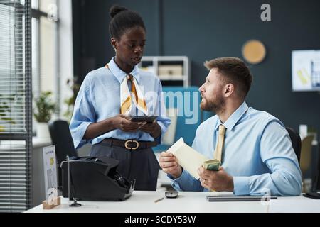 Jeune femme noire debout à côté d'un homme caucasien assis à un bureau, femme tenant une calculatrice et un bloc-notes, homme comptant l'argent comptant et tenant une enveloppe dans un bureau de banque moderne Banque D'Images