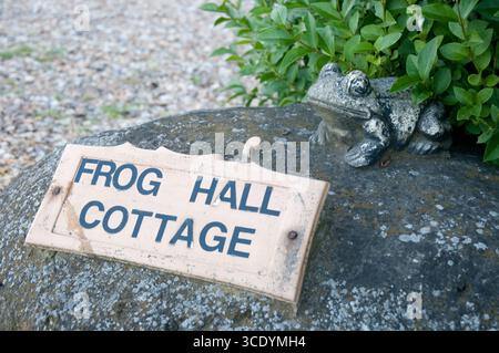 Un charmant signe pour Frog Hall Cottage se trouve sur une pierre, accompagné d'une grenouille décorative au milieu du feuillage vert dans un jardin serein. Banque D'Images