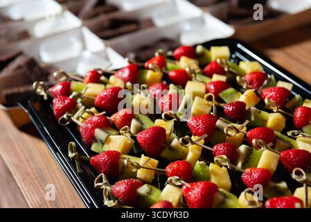 Une exposition vibrante avec des brochettes de fraises fraîches, kiwi et ananas, accompagnés de desserts au chocolat en arrière-plan. parfait pour faire la fête Banque D'Images