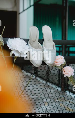 Une paire de pantoufles à talons hauts en argent pailleté accrochées à une clôture en métal noir, ornée de fleurs blanches et roses. L'image crée un beau et Banque D'Images