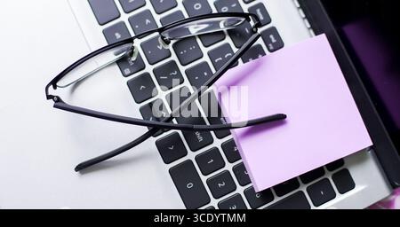 Les lunettes reposent sur un clavier d'ordinateur portable à côté d'une note autocollante, créant une atmosphère de bureau à domicile confortable. Cette configuration suggère un environnement de travail actif rempli Banque D'Images