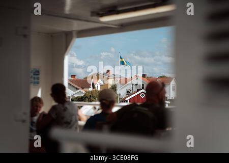Brännö, Göteborgs Schärengarten, Schweden Blick von einer Fähre auf rote, weiße und gelbe Holzhäuser auf Brännö, schwedische Flagge im Wind, unscharfe Passagiere im Vordergrund. Brännö Västra Götalands län Brännö Schweden *** Brännö, archipel de Gothenburg, Suède vue d'un ferry sur des maisons en bois rouges, blanches et jaunes sur Brännö, drapeau suédois dans le vent, passagers flous au premier plan Brännö Västra Götalands län Brännö Suède Banque D'Images