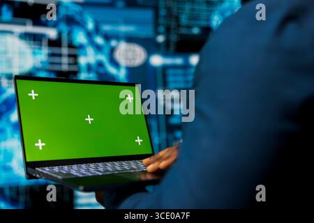 Jeune spécialiste masculin examine les données algorithmiques à côté de l'écran vert, utilise un ordinateur portable entouré de visuels numériques. La technologie intelligente DE L'IA et les réseaux neuronaux dans un environnement de haute technologie. Banque D'Images