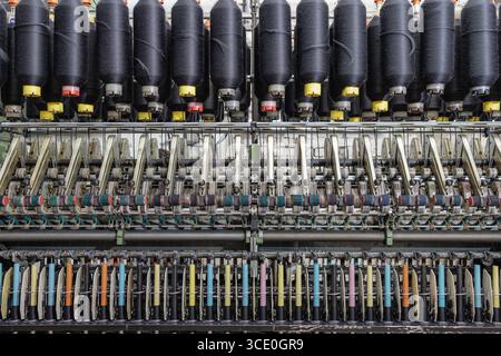 Vue de face d'un métier à filer automatisé avec des cônes de fil noir et des broches dans une usine textile, montrant la production moderne. Banque D'Images