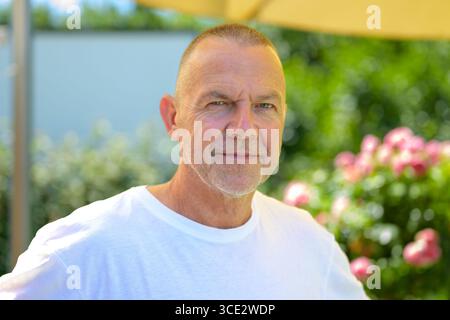Portrait en gros plan d'un homme âgé confiant portant un t-shirt blanc, debout dans un jardin ensoleillé avec des fleurs roses en arrière-plan. Banque D'Images