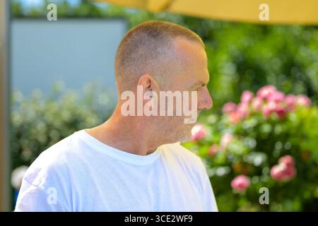 Portrait de profil de côté d'un homme mature dans un t-shirt blanc, debout à l'extérieur devant un jardin fleuri. Banque D'Images