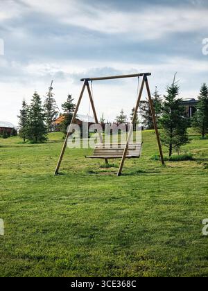 Balançoire vide dans le parc. Balançoires en bois sur une grande pelouse, plantes à feuilles persistantes en arrière-plan. Banque D'Images