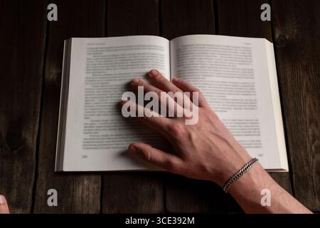 Un gros plan d'une main ornée d'un bracelet en argent reposant sur les pages ouvertes d'un livre sur une table en bois rustique, évoquant un guichet automatique confortable de lecture et d'étude Banque D'Images