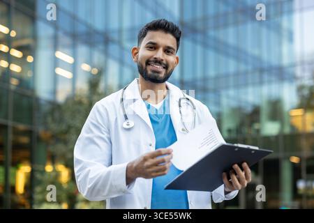 Un médecin joyeux avec stéthoscope autour du cou, tenant une planche à pince, posant à l'extérieur avec confiance et positivité. Banque D'Images