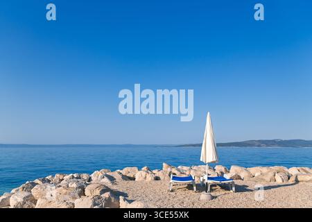 Scène estivale minimaliste : deux chaises de plage vides et un parasol sur une côte rocheuse avec une mer bleue calme et un ciel dégagé. Vacances parfaites en Croatie Banque D'Images