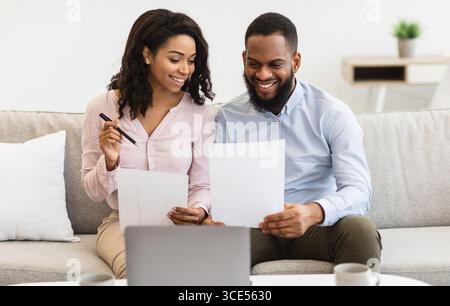 Couple noir joyeux lisant des documents à la maison, vérifiant des factures Banque D'Images