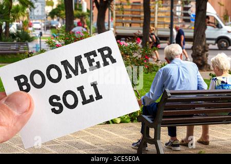 Bavière, Allemagne - 13 août 2025 : MONTAGE PHOTO, Une main tient une carte avec les mots Boomer Soli écrits dessus, tandis que des personnes âgées sont assises sur un banc de parc en arrière-plan, symbole des discussions politiques sur le financement des pensions et la solidarité intergénérationnelle. *** FOTOMONTAGE, Eine Hand hält eine Karte mit der Aufschrift Boomer Soli, im Hintergrund sitzen ältere Menschen auf einer Parkbank Sinnbild für politische Diskussionen um Rentenfinanzierung und Generationensolidarität. Banque D'Images