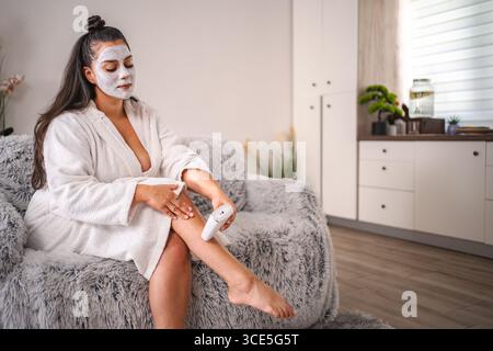 Jeune femme enlevant les poils de jambe tout en portant un masque facial à la maison Banque D'Images