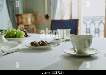 Gros plan montrant tasse en porcelaine et soucoupe sur la table avec assiette de raisins verts et rouges, bol de biscuits au chocolat, et tasse dans une pièce élégante avec la lumière du soleil coulant par la fenêtre Banque D'Images