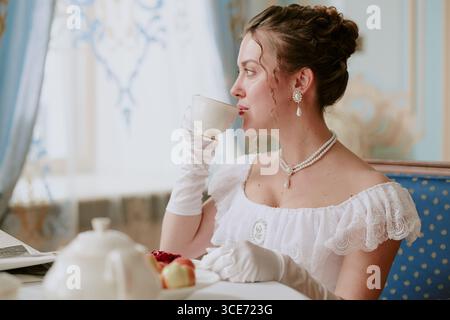 Femme jeune adulte caucasienne assise à table buvant de la tasse de thé portant des bijoux et des gants élégants, regardant à côté avec les cheveux coiffés, appréciant la mise à thé raffinée Banque D'Images
