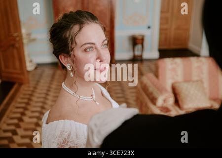Femme jeune adulte caucasienne avec des cheveux bruns bouclés portant des bijoux en perles souriant et regardant vers le haut, tout en se tenant à l'intérieur près de l'homme dans une tenue formelle lors d'un événement élégant Banque D'Images