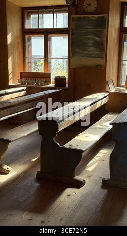 De vieux bureaux et bancs en bois dans une salle de classe d'école de village rustique évoquent un sentiment de nostalgie. La lumière du soleil filtre à travers les grandes fenêtres, mettant en évidence le Banque D'Images