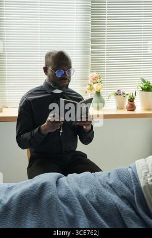 Aumônier mâle noir priant et lisant la Bible à côté du lit d'hôpital Banque D'Images
