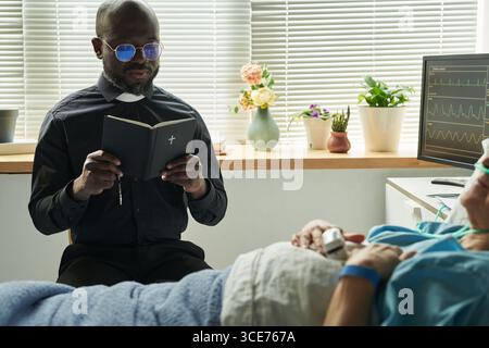 Prêtre noir d'âge moyen priant aux côtés d'une femme caucasienne senior hospitalisée Banque D'Images