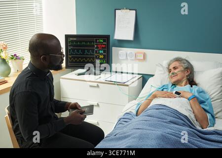 Femme caucasienne d'âge moyen allongée dans le lit d'hôpital parlant au prêtre noir Banque D'Images