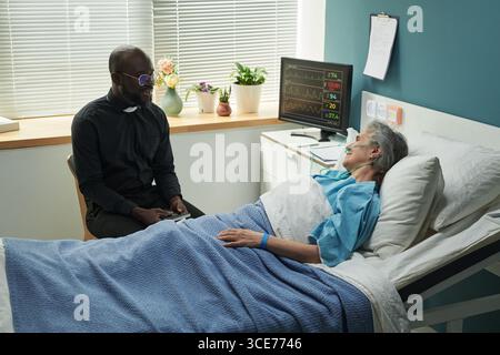 Prêtre mâle noir d'âge moyen réconfortant la femme caucasienne senior dans le lit d'hôpital Banque D'Images