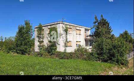 La maison blanche vieillie présente des fenêtres bordées et une façade pelable, entourée de verdure luxuriante, de buissons et d'arbres contre un ciel bleu vif un jour d'été Banque D'Images
