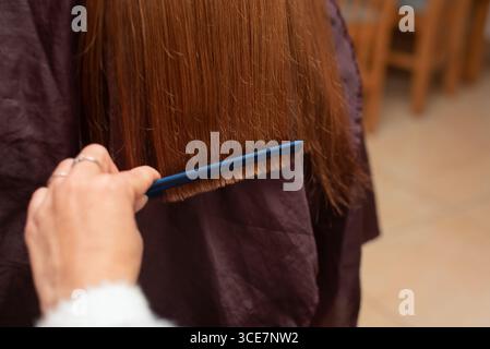 Vue rapprochée montrant des mains peignant soigneusement les cheveux bruns avec un peigne bleu dans un salon de coiffure, mettant en évidence les pratiques de toilettage et d'hygiène Banque D'Images