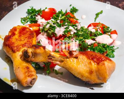 Deux appétissantes cuisses de poulet grillé sont placées sur une assiette de couleur claire à côté d'une salade fraîche de tomates tranchées, de roquette et d'un d crémeux à base de yaourt Banque D'Images