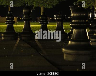 Un cliché saisissant en bas angle de grandes pièces d'échecs sombres sur un plateau de jeu carrelé dans un parc la nuit. Banque D'Images