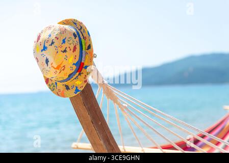 Chapeau de paille coloré reposant sur un poteau de hamac en bois avec mer turquoise en arrière-plan, parfait pour les thèmes de voyage d'été Banque D'Images