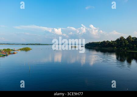 Vue tranquille sur le lac Kaptai avec une rive boisée verdoyante, des plantes aquatiques dispersées et un ciel bleu serein Banque D'Images