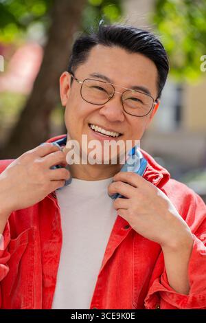 Portrait d'homme d'âge moyen asiatique souriant heureux écoutant de la musique décollant des écouteurs regardant la caméra, la détente se sentir satisfait de bonnes nouvelles en plein air. Mec mûr chinois sur la rue de la ville urbaine. Vertical Banque D'Images